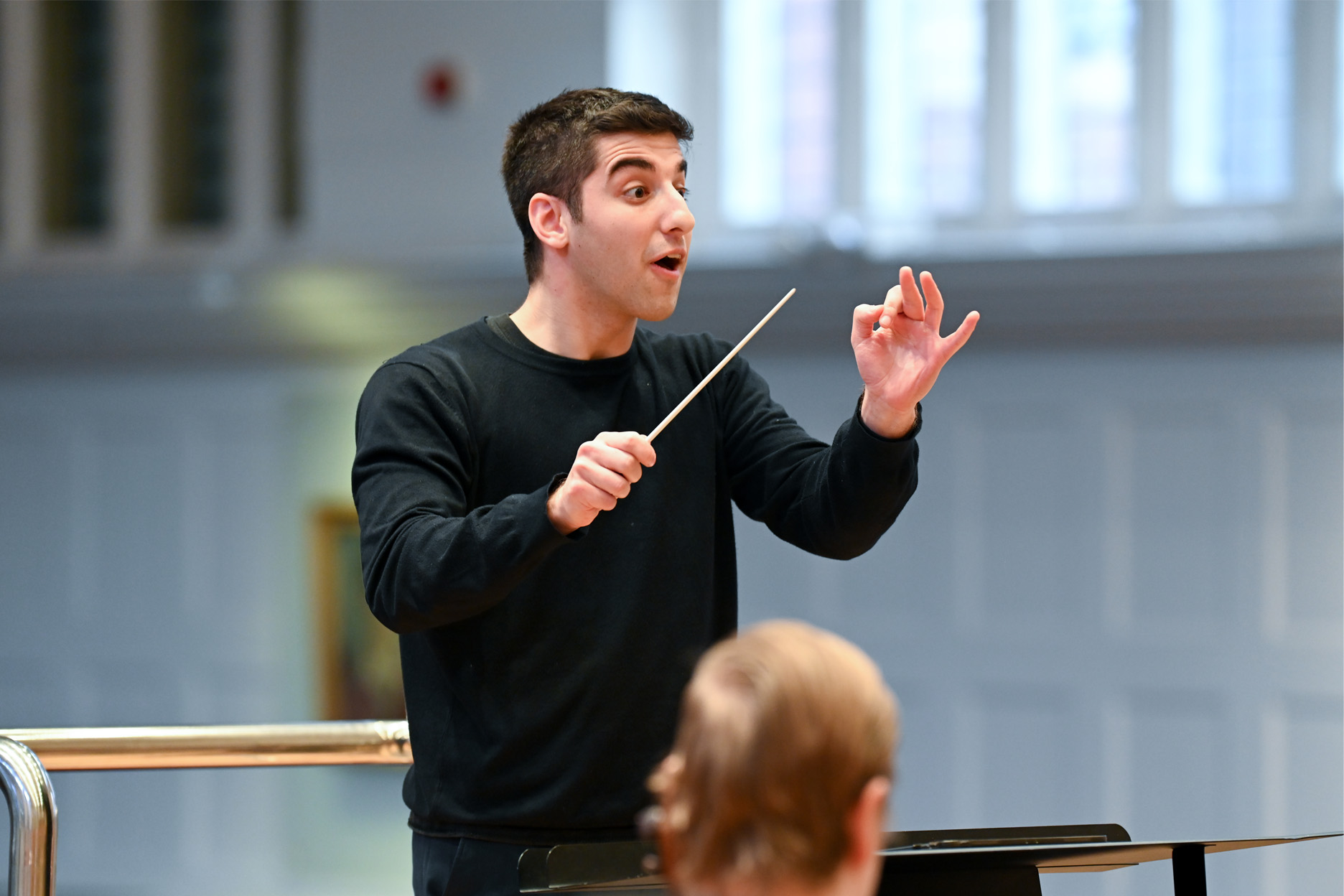 Male student, conducting a rehearsal for an upcoming orchestra, with musicians in the foreground in RCM's Amaryllis Fleming Concert Hall